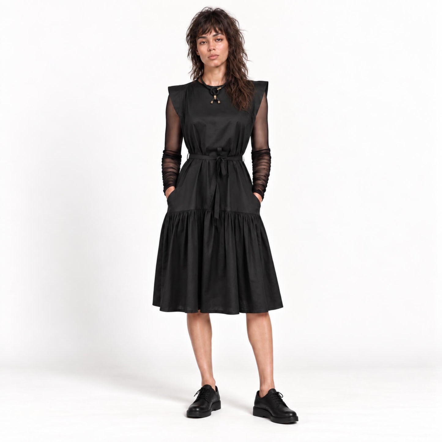 A woman modeling a black midi dress with sheer long sleeves, a tie waist, and a tiered skirt, paired with black shoes against a white studio background.