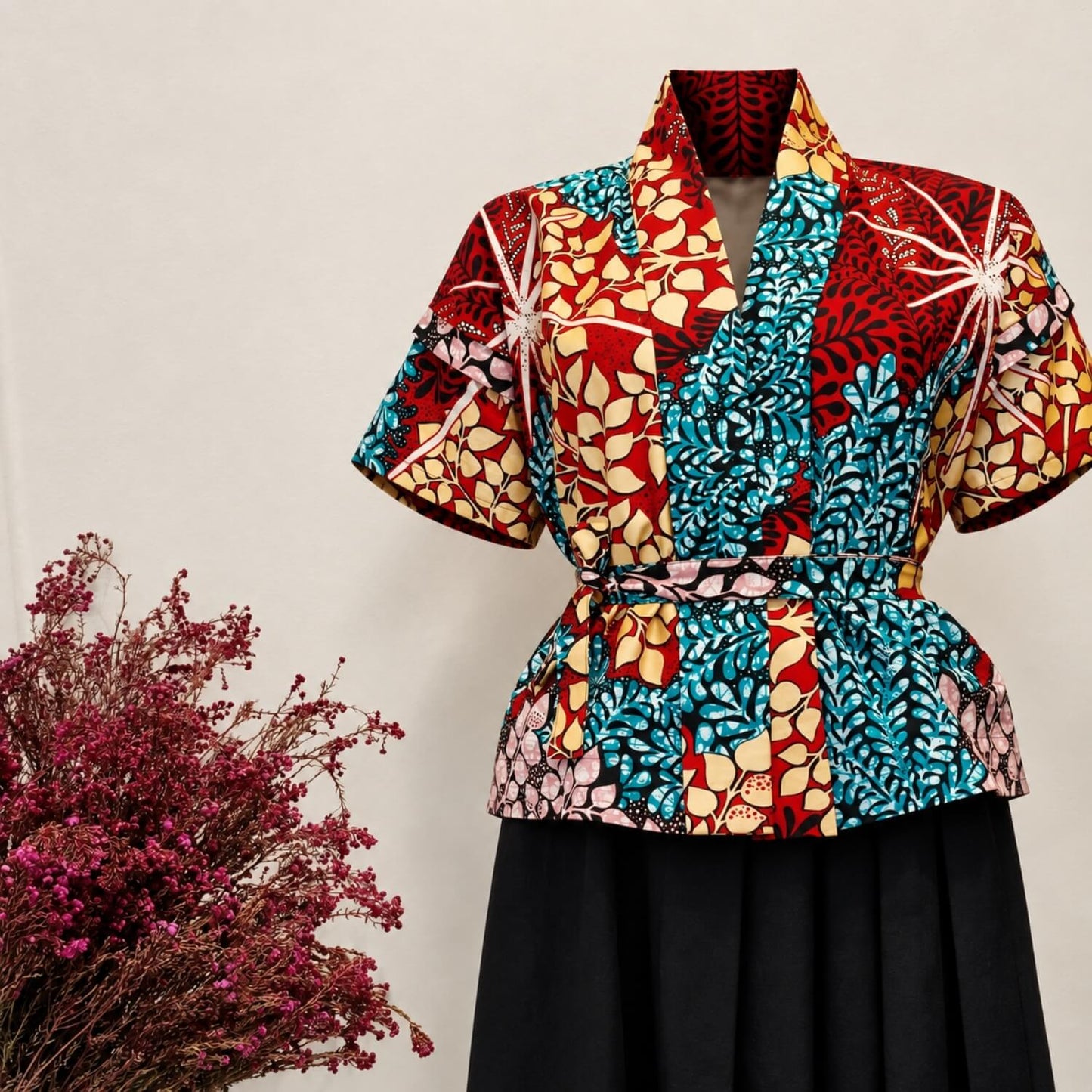 A short-sleeve belted wrap top with a bold multicolor floral and geometric print in red, turquoise, cream, and pink, styled on a mannequin with a black skirt against a light beige background with dried pink flowers at the side.
