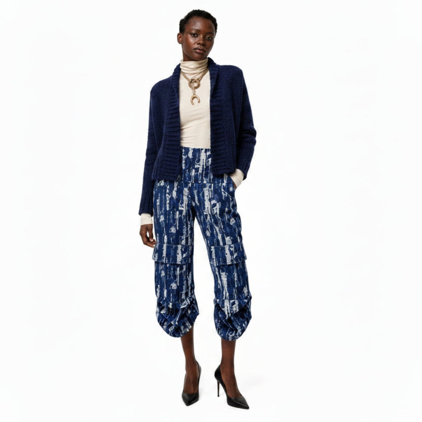 A model stands against a white studio background wearing a navy blue textured cardigan, a cream turtleneck, and navy cargo pants with a white distressed pattern.