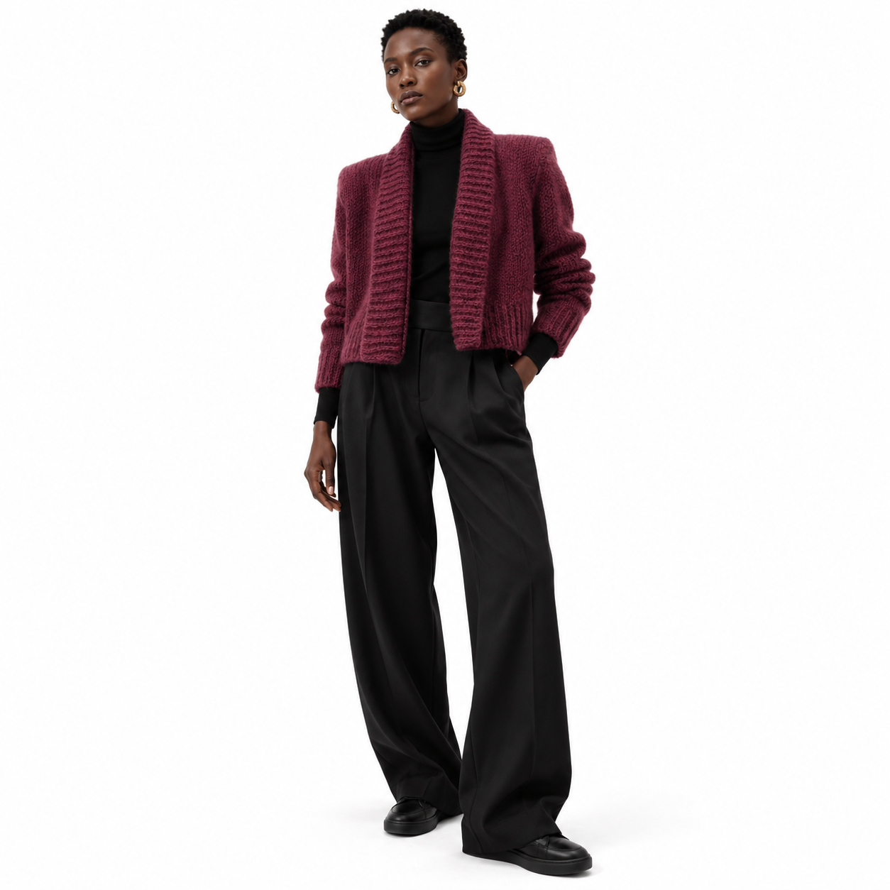 A model stands against a white studio background wearing a textured burgundy cropped cardigan over a black turtleneck, paired with black wide-leg trousers and black shoes.