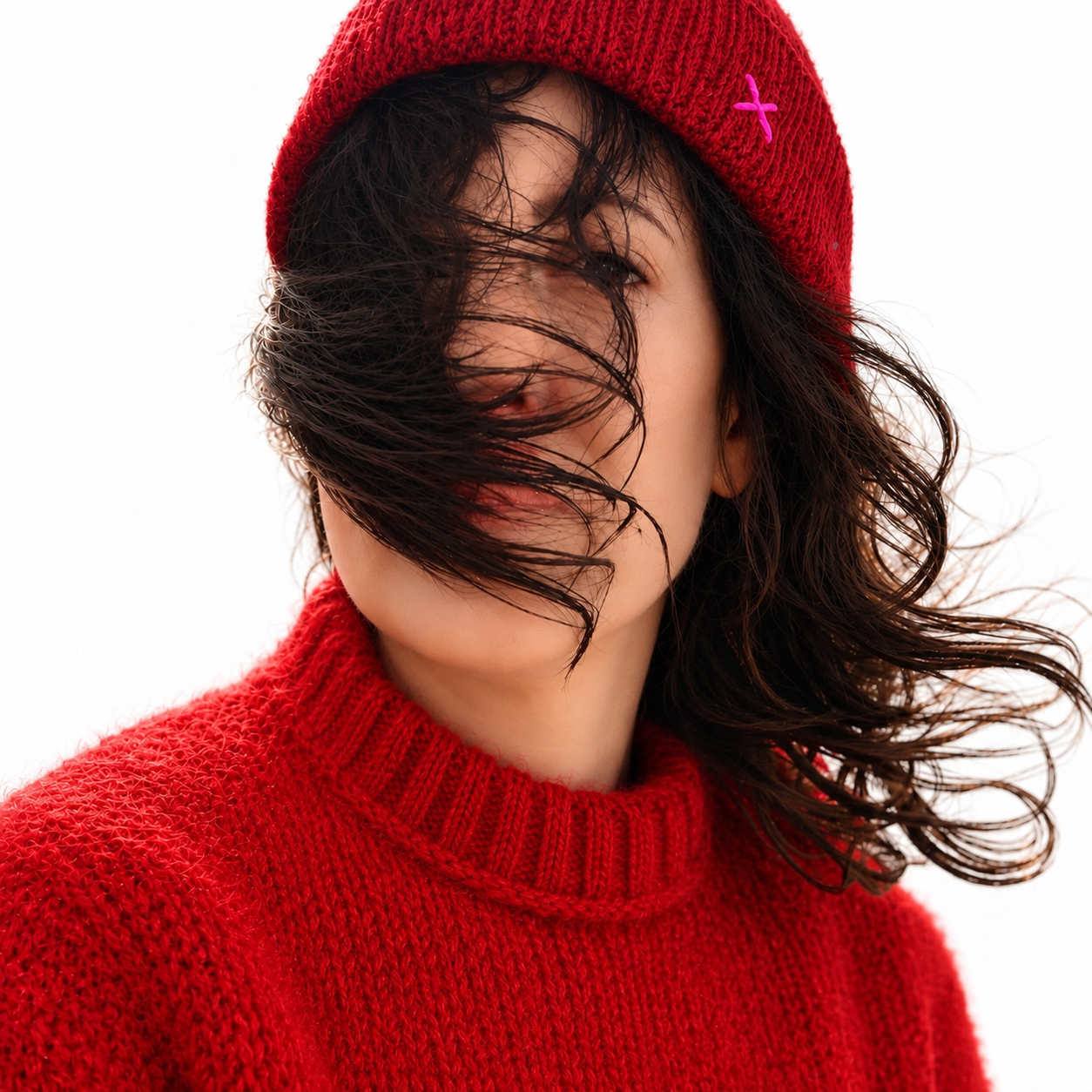 A close-up portrait of a woman wearing a bright red knit beanie with a small pink cross detail and a matching red knit sweater, with her dark hair blowing across her face against a white background.