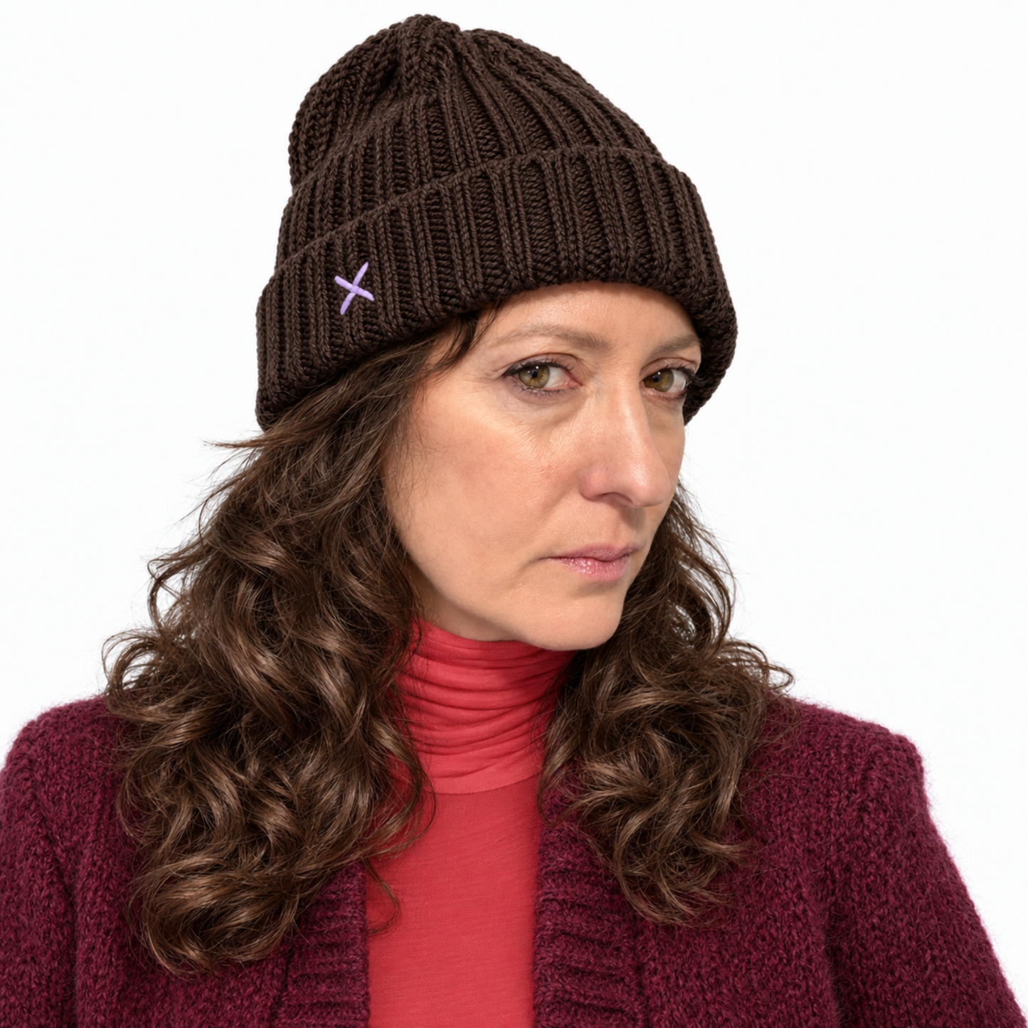 A woman wearing a dark brown ribbed knit beanie with a small purple embroidered cross on the side, paired with a red turtleneck and burgundy cardigan, against a plain white background.