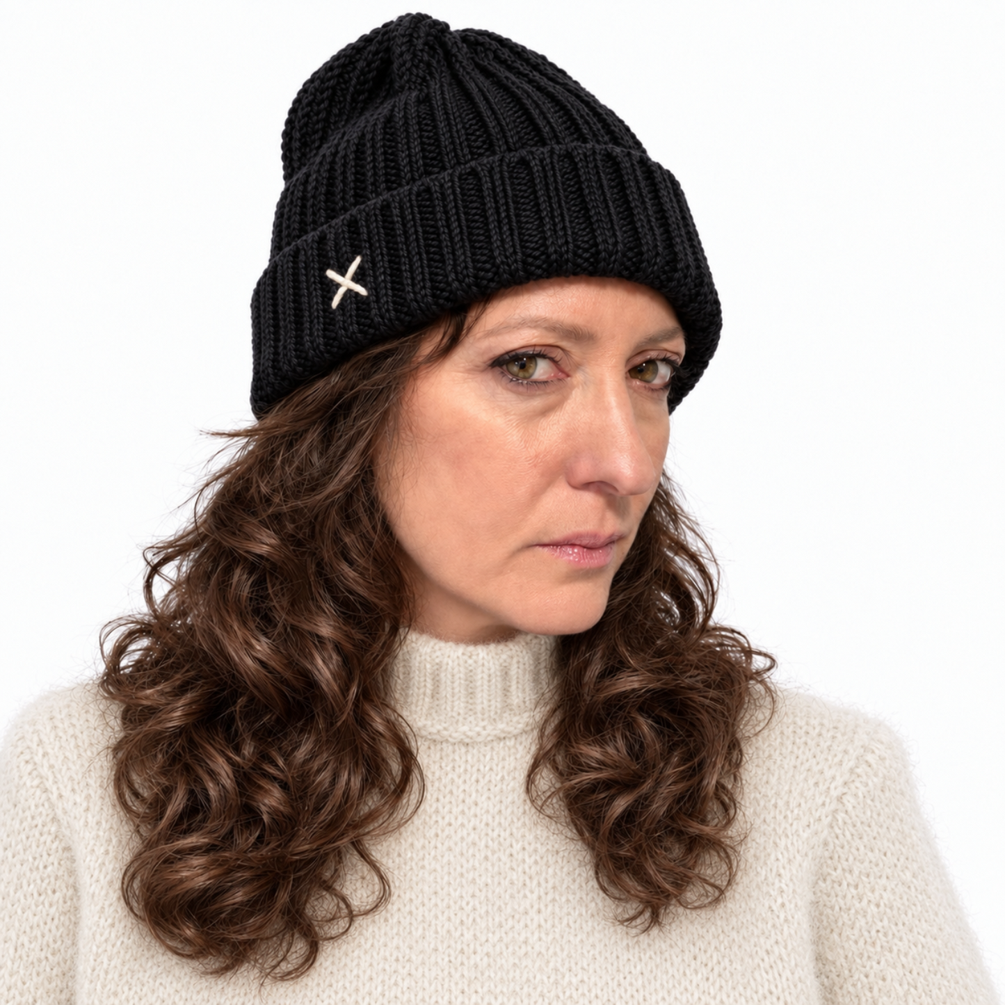 A woman wearing a black ribbed knit beanie with a small white embroidered cross on the side, paired with a cream knit sweater, against a plain white background.