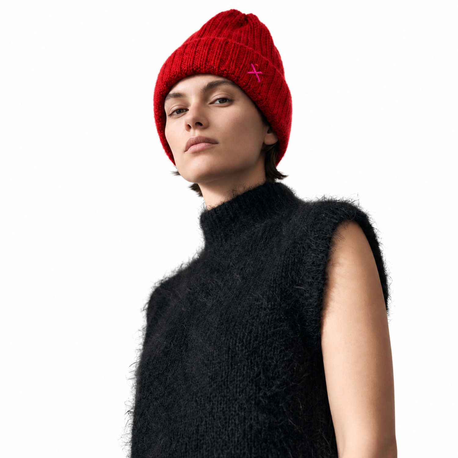 A model wearing a bright red ribbed knit beanie with a small pink embroidered cross on the side, paired with a fuzzy black sleeveless mock-neck sweater, against a solid white background.