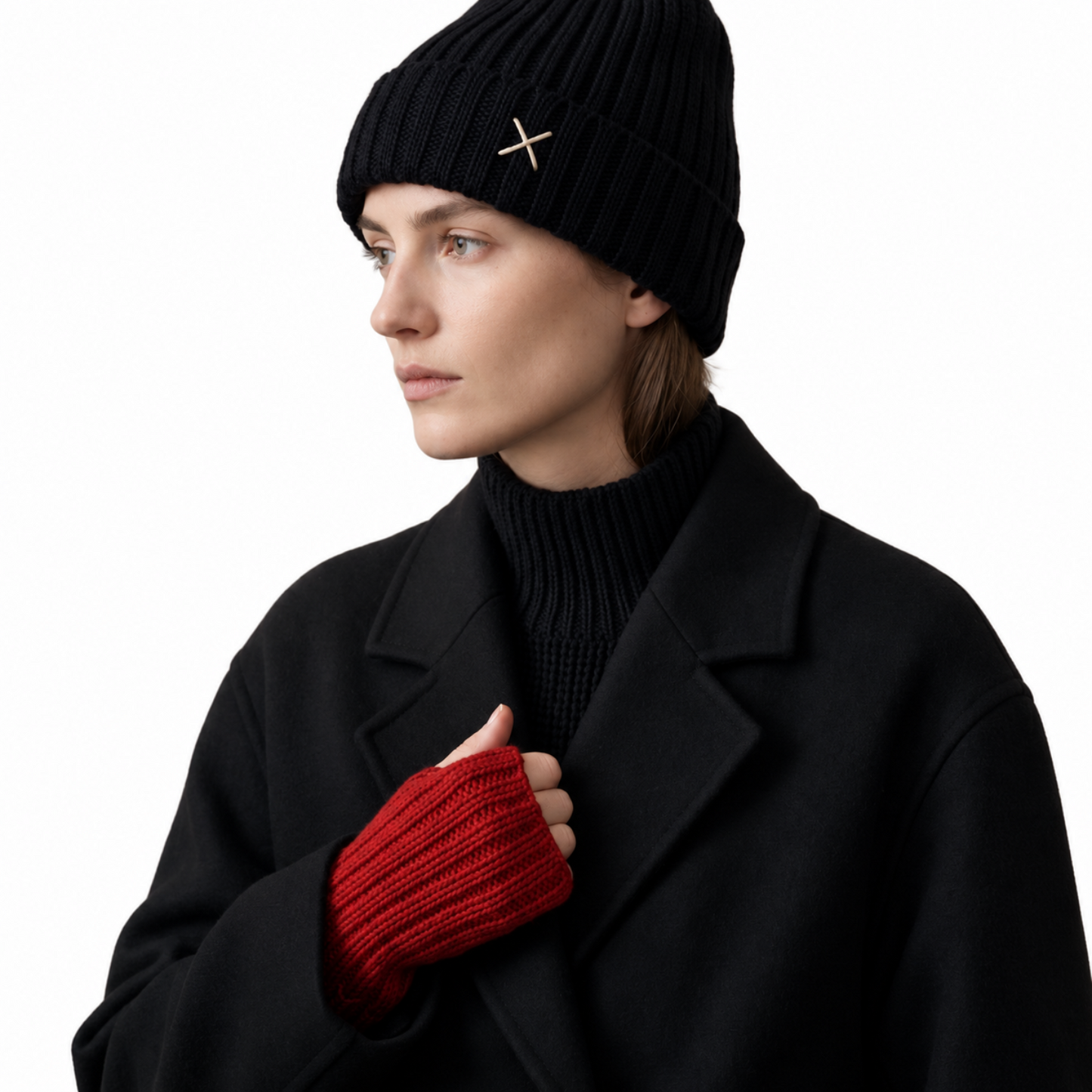 A woman wearing a black ribbed knit beanie with a small gold cross emblem, a black turtleneck, a black coat, and a red ribbed knit fingerless glove, shown against a plain white background.