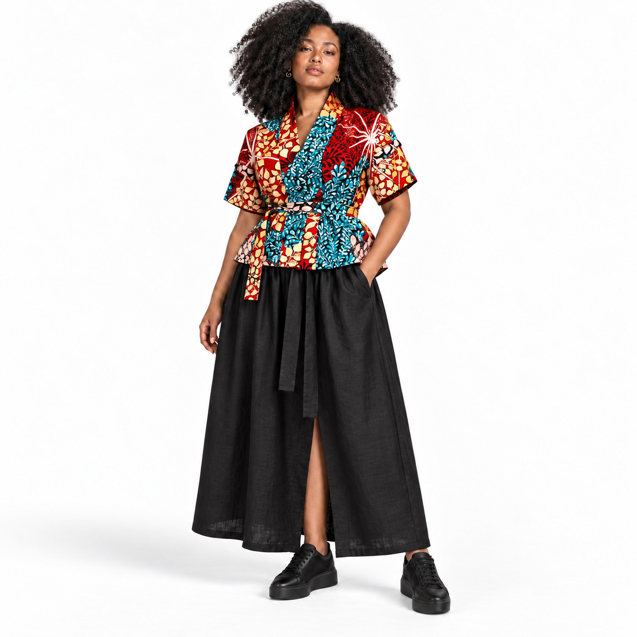 A model with curly black hair is wearing a short-sleeve wrap-style top with a vibrant multicolor African wax print (red, blue, orange, yellow) paired with a long black skirt and black platform sneakers, shot against a solid white studio background.