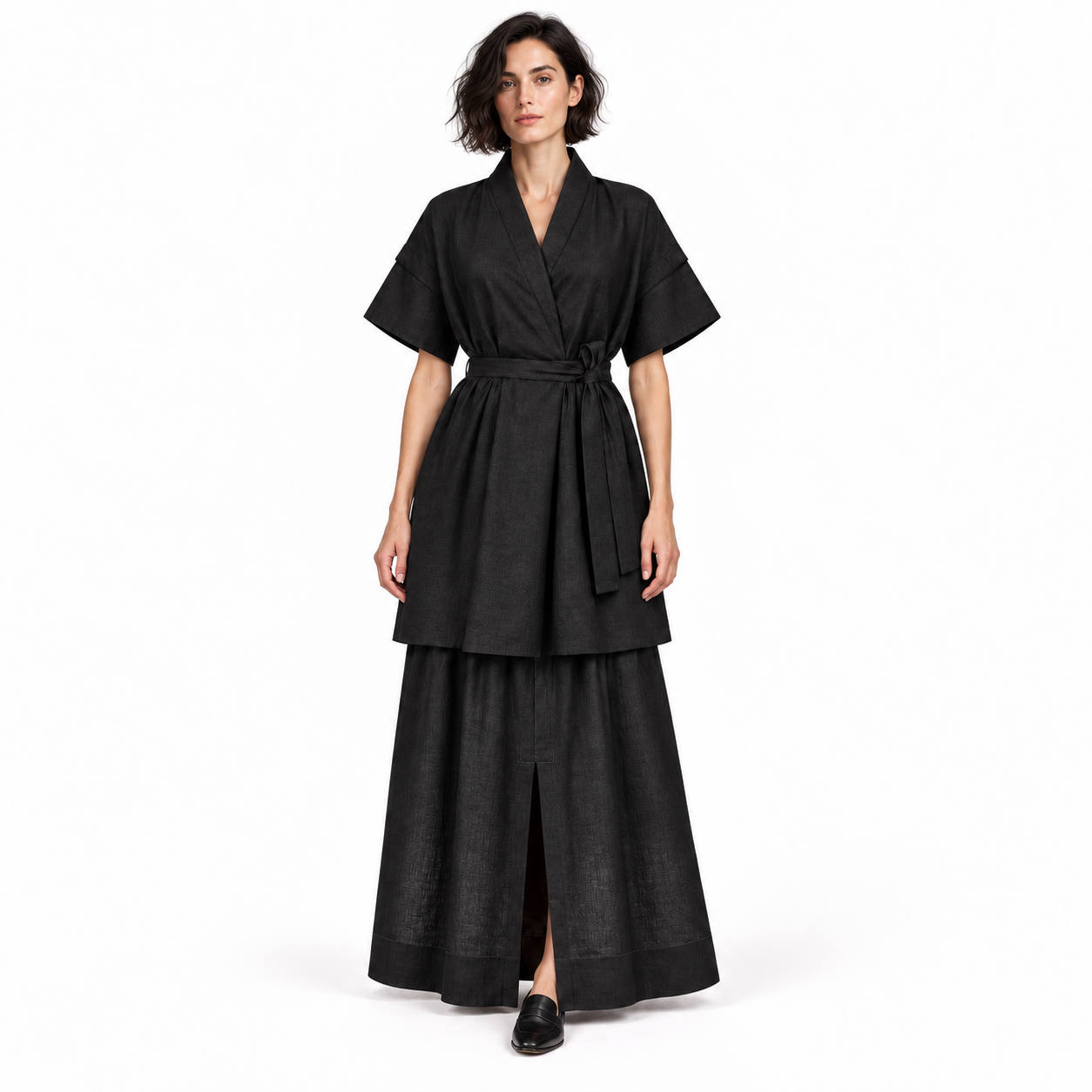 A woman modeling a black two-piece linen outfit consisting of a short-sleeve belted wrap top and a matching high-waisted maxi skirt, photographed against a solid white studio background.