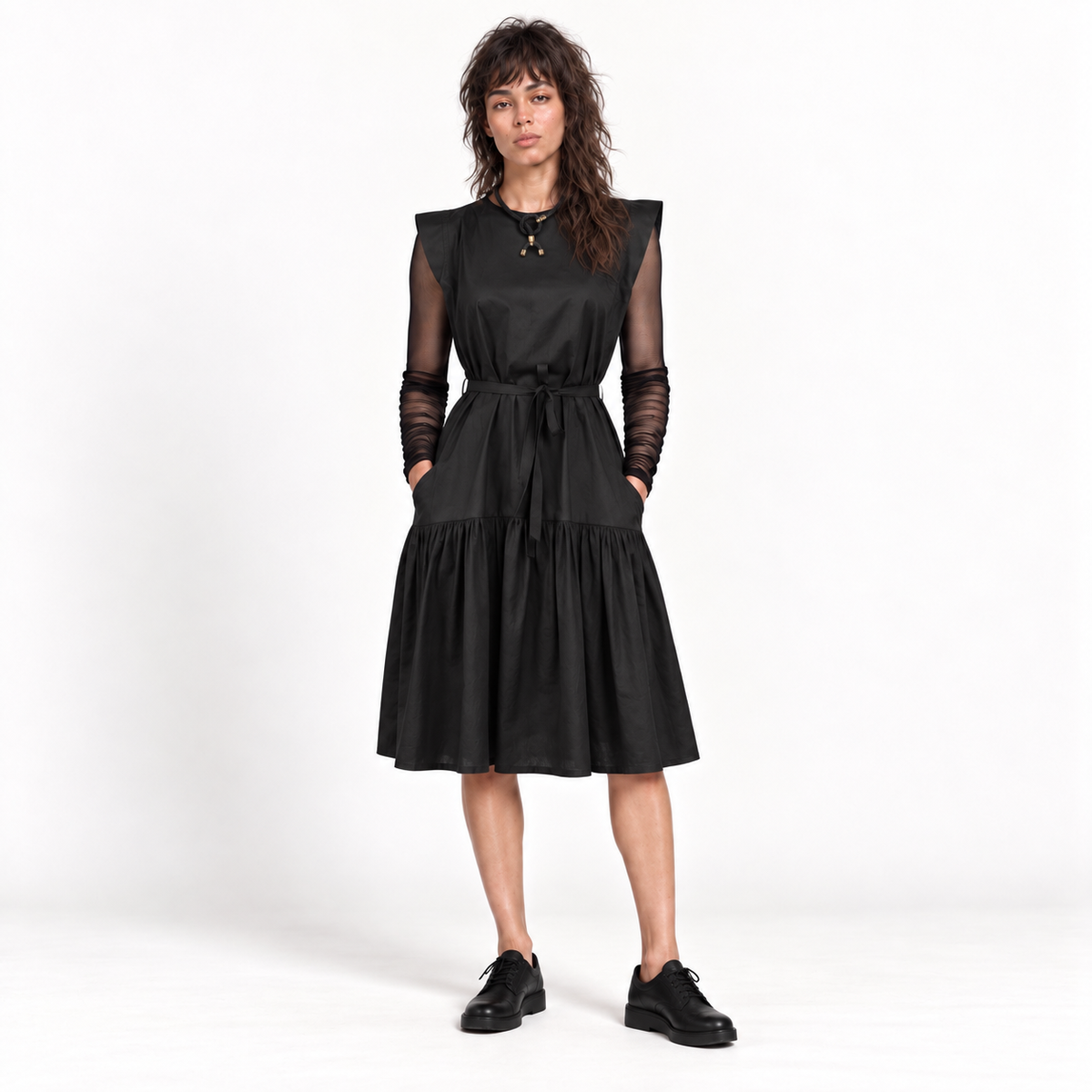 A woman modeling a black midi dress with sheer long sleeves, a tie waist, and a tiered skirt, paired with black shoes against a white studio background.