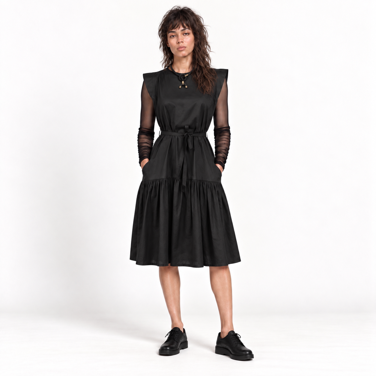 A woman modeling a black midi dress with sheer long sleeves, a tie waist, and a tiered skirt, paired with black shoes against a white studio background.