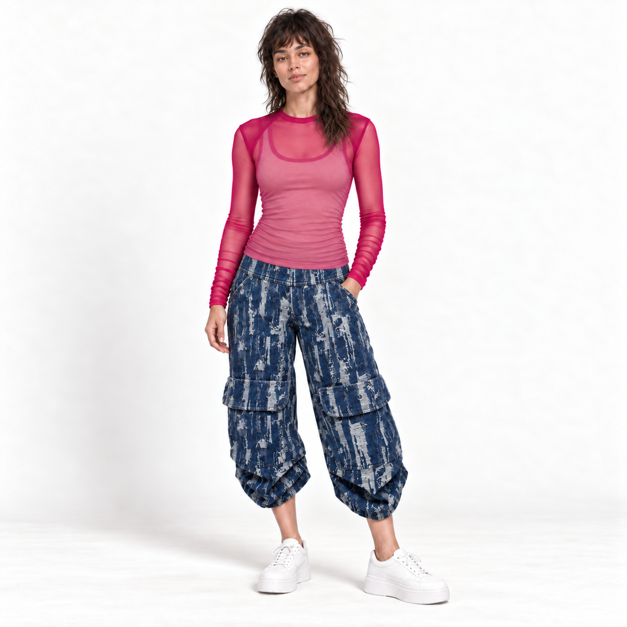 A model stands against a white studio background wearing a pink long-sleeve mesh top, blue-and-white patterned cargo pants, and white sneakers.