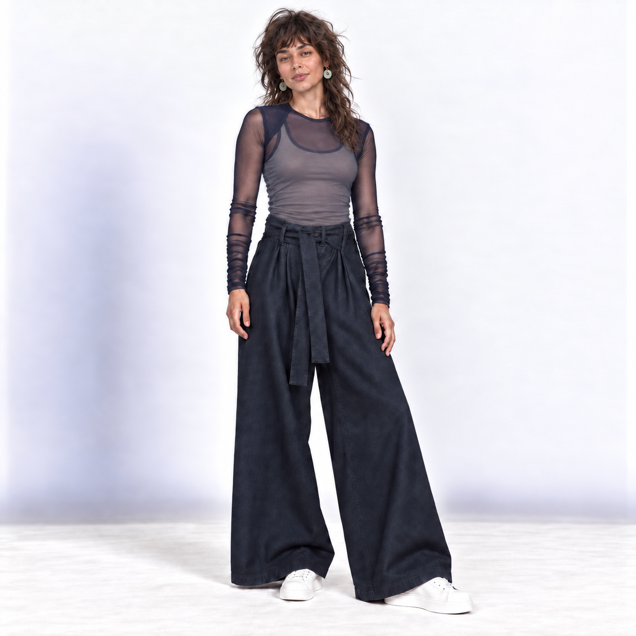 A woman modeling a dark gray sheer long-sleeve mesh top with black lace-up cuffs, paired with dark navy wide-leg pants and white sneakers, against a light gray studio backdrop.