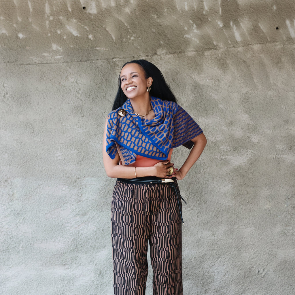 A woman posing in front of a textured concrete wall, wearing a blue and orange patterned wrap-style top and matching patterned trousers.