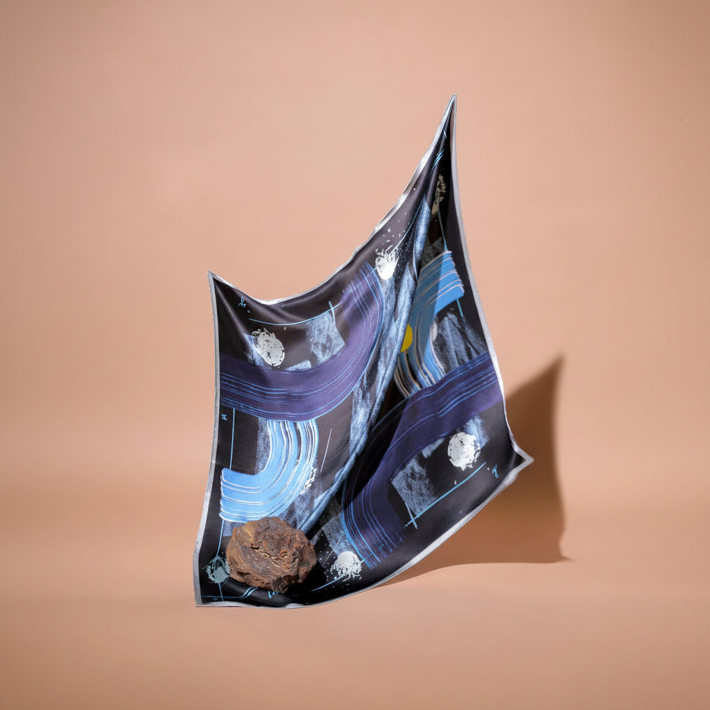 A square scarf with an abstract space-themed print featuring black and blue brushstrokes, white celestial bodies, and a small yellow sun on a black background, draped dynamically against a solid peach background.