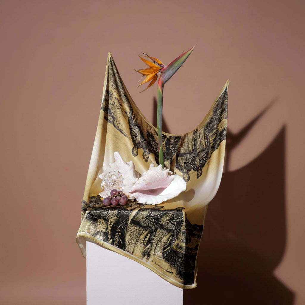 A draped scarf with a black and gold bird pattern, styled with seashells, purple grapes, and an orange bird of paradise flower against a solid brown background.