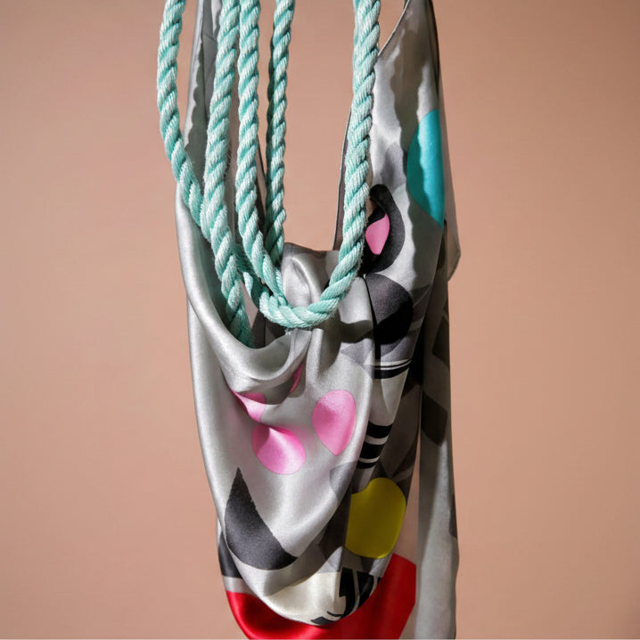 A close-up of a satin scarf with a gray, black, and pink abstract pattern, featuring thick twisted rope handles in mint green.
