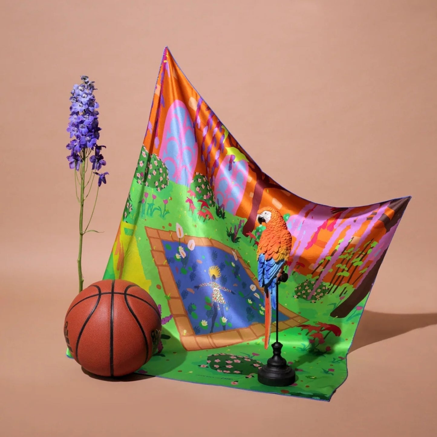 A colorful printed fabric panel or scarf-like sheet draped on a beige studio background, featuring a painted pond scene with a parrot, trees, and bright geometric patterns. A basketball and a purple flower stem are placed beside it as styling props.