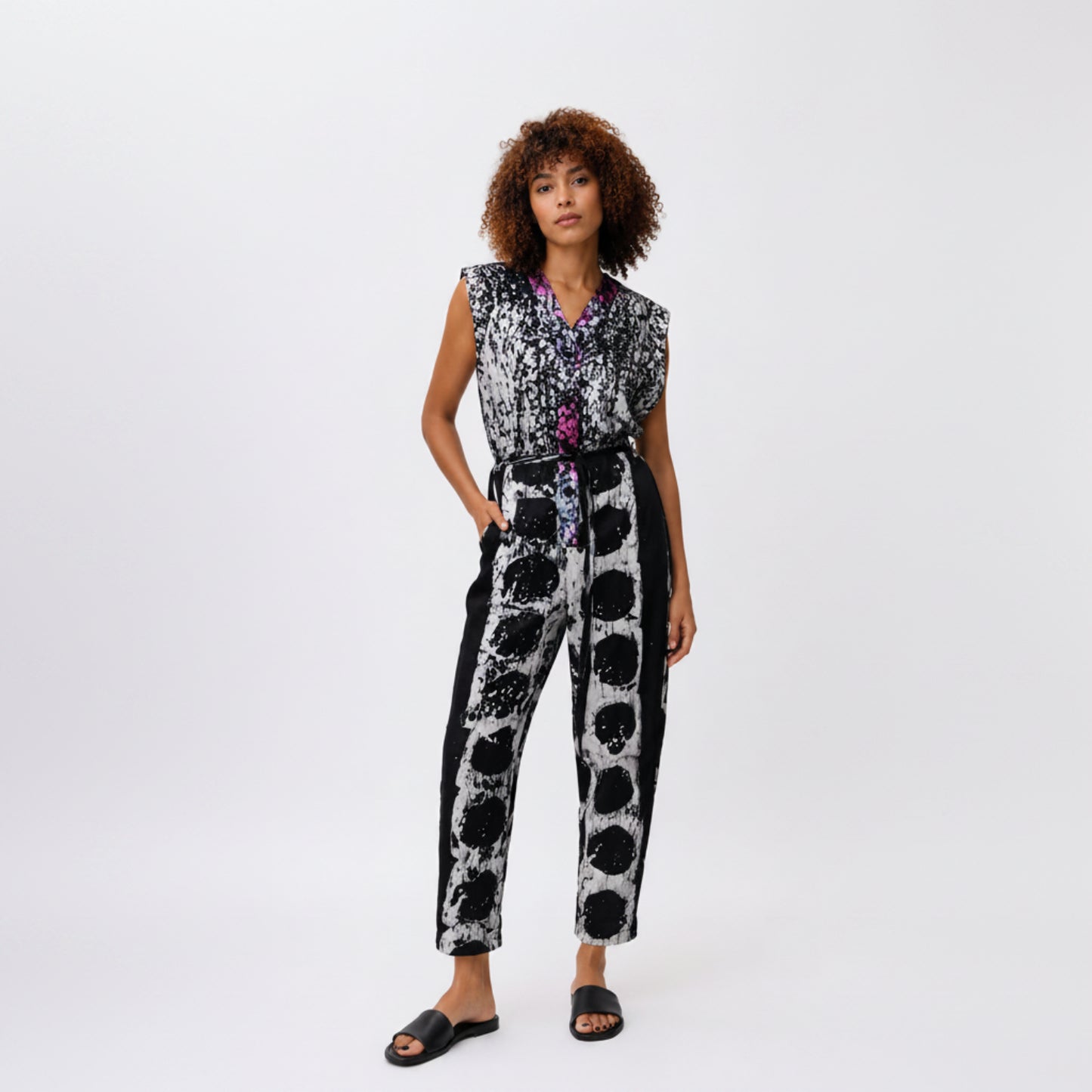 A model wearing a sleeveless black and white patterned jumpsuit with a purple and blue patterned inner collar, paired with black slide sandals, against a plain white studio background.
