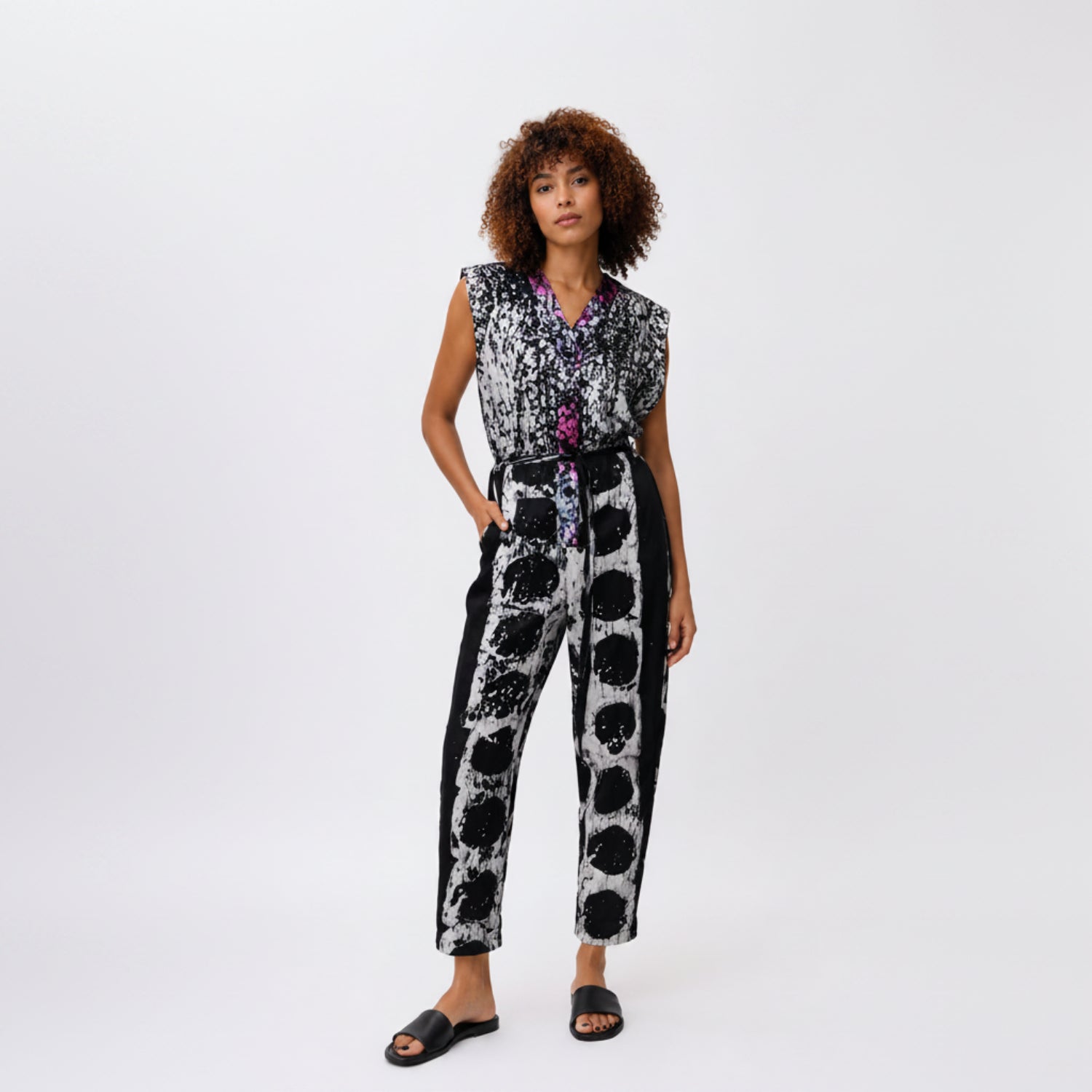 A model wearing a sleeveless black and white patterned jumpsuit with a purple and blue patterned inner collar, paired with black slide sandals, against a plain white studio background.