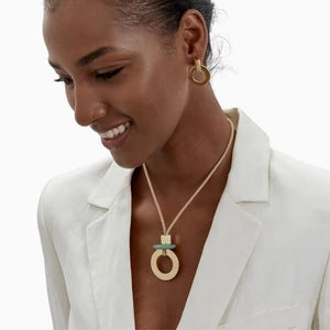 A woman wearing a gold pendant necklace with a circular ring and a small turquoise accent, styled with matching gold hoop earrings and a white blazer.