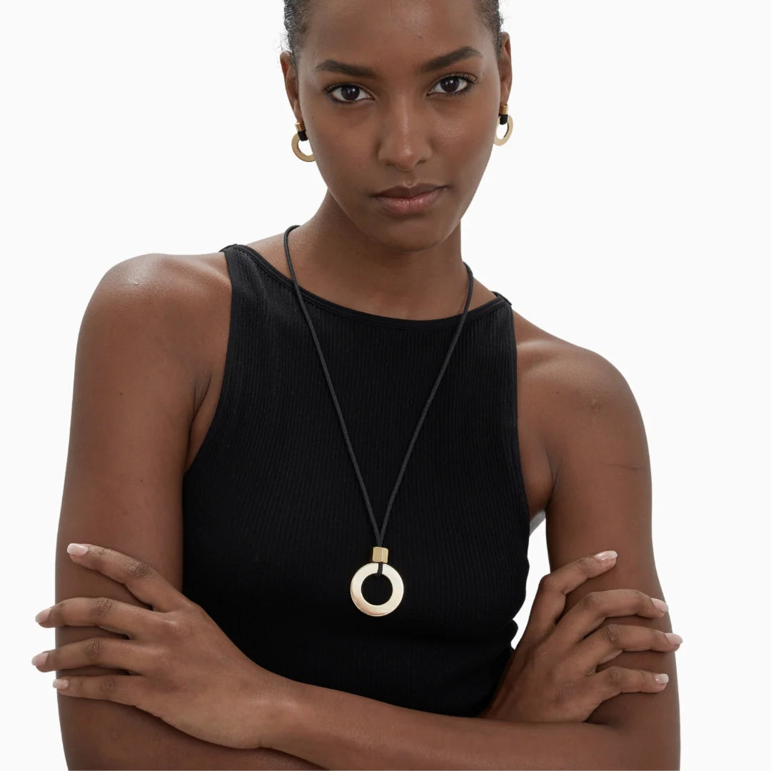 A woman wearing a black ribbed sleeveless top and a long black cord necklace with a gold-toned circular pendant, photographed against a plain white background.
