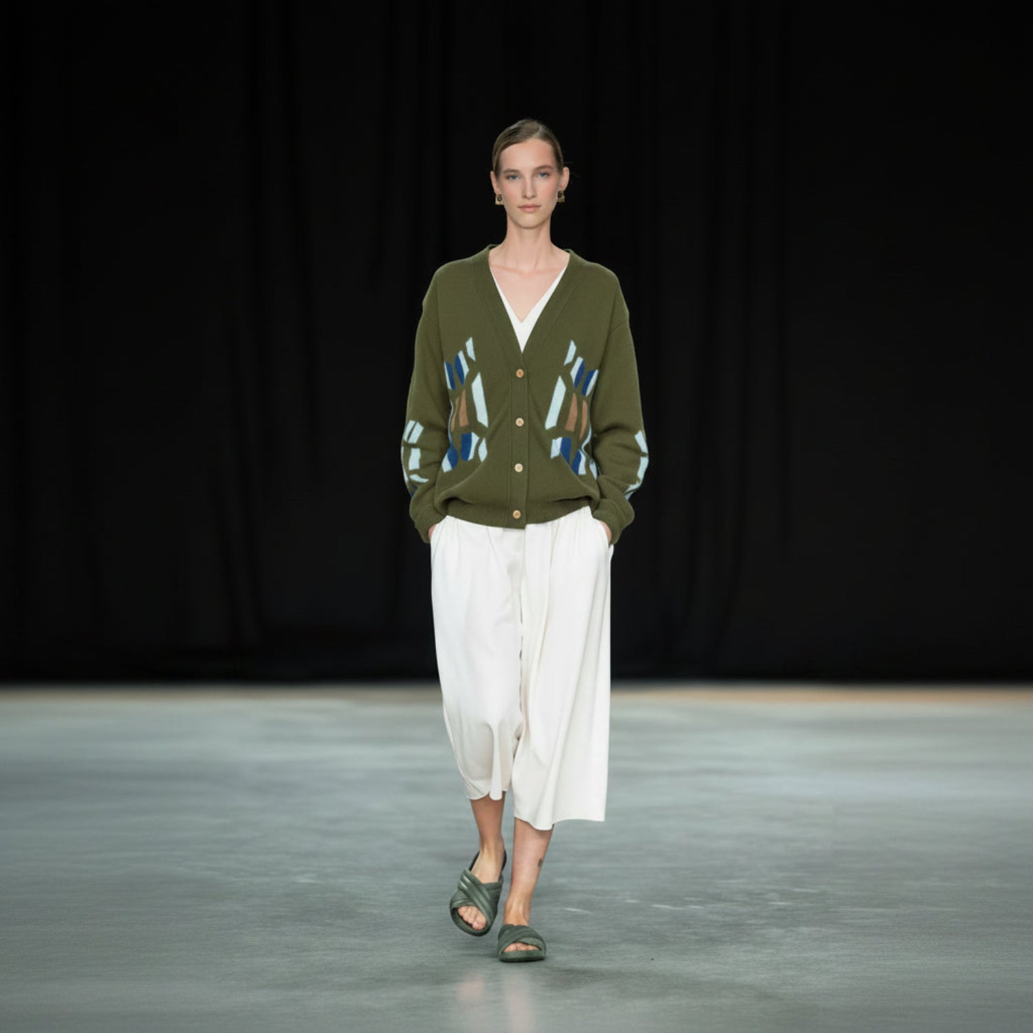 A model walks a runway wearing an olive green cardigan with abstract blue, brown, and white knit patterns on the front and sleeves, paired with a white midi skirt and green sandals.