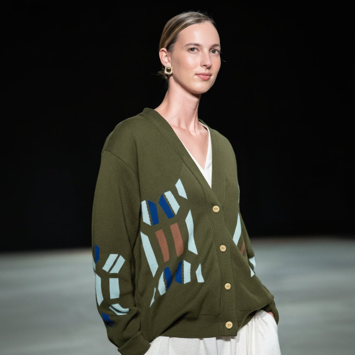 A model on a runway wearing an olive green knit cardigan with geometric blue, beige, and brown patterns on the front, paired with a white V-neck undershirt and a gold geometric earring.