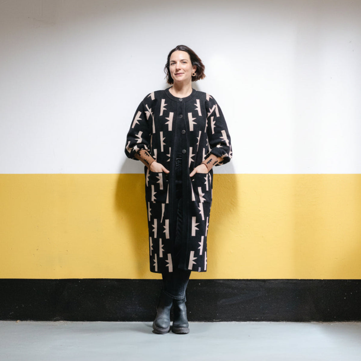 A woman stands against a two-tone wall painted white on the upper half and yellow on the lower half, wearing a long black coat with a repeating beige rifle pattern, black pants, and black boots.
