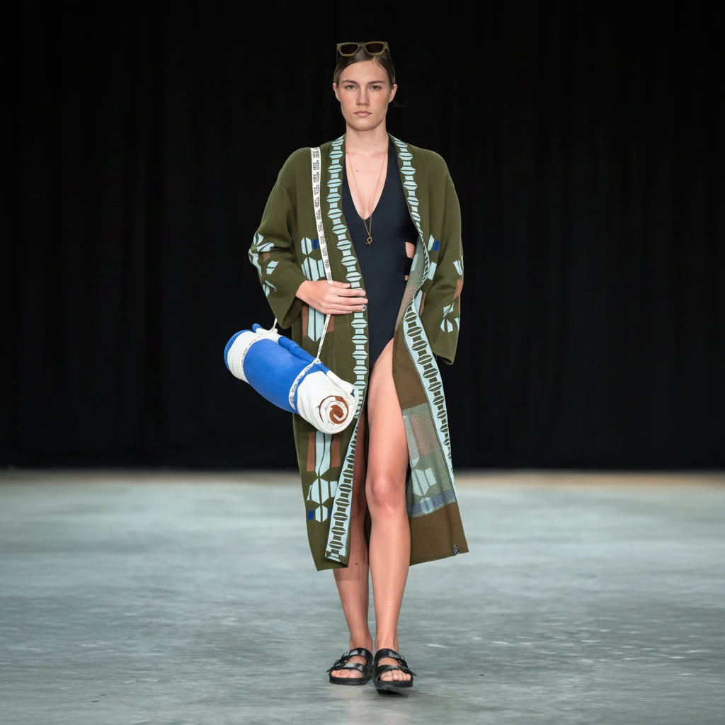 A model walks a runway wearing an olive green open-front cardigan with a light blue and white geometric pattern, layered over a black swimsuit. She is holding a rolled-up blue and white towel or blanket against a solid black background.