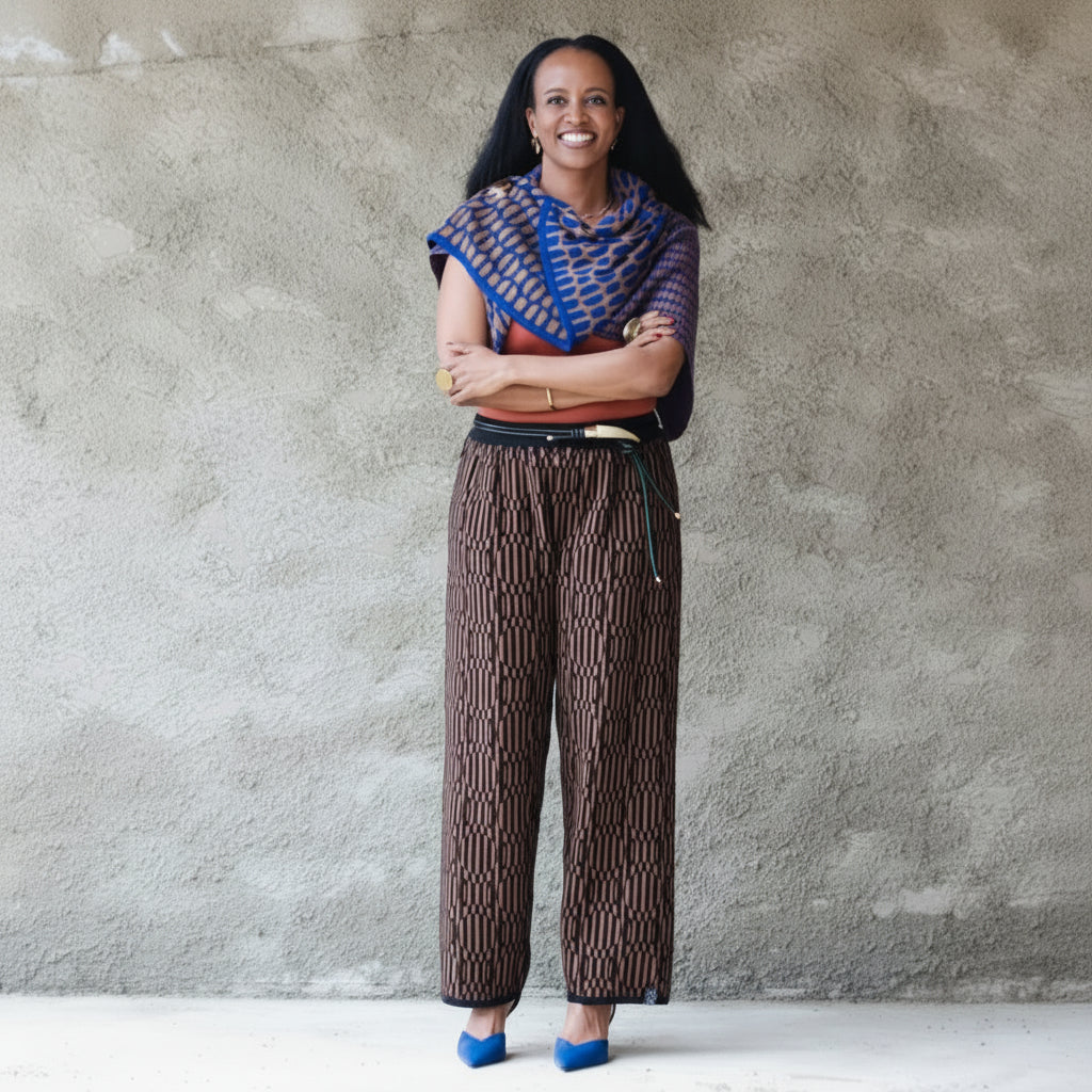 A woman stands against a textured gray concrete wall wearing a vibrant blue and brown patterned shawl draped over her shoulders, paired with brown patterned wide-leg trousers and bright blue high heels.
