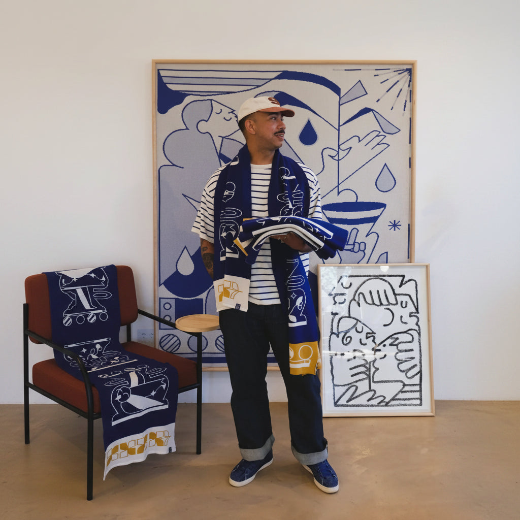 A man wearing a navy and white patterned knit scarf, striped shirt, and dark pants stands in a room with framed abstract art. The scarf has a blue and white graphic pattern with yellow accents, and matching scarves are draped over furniture.
