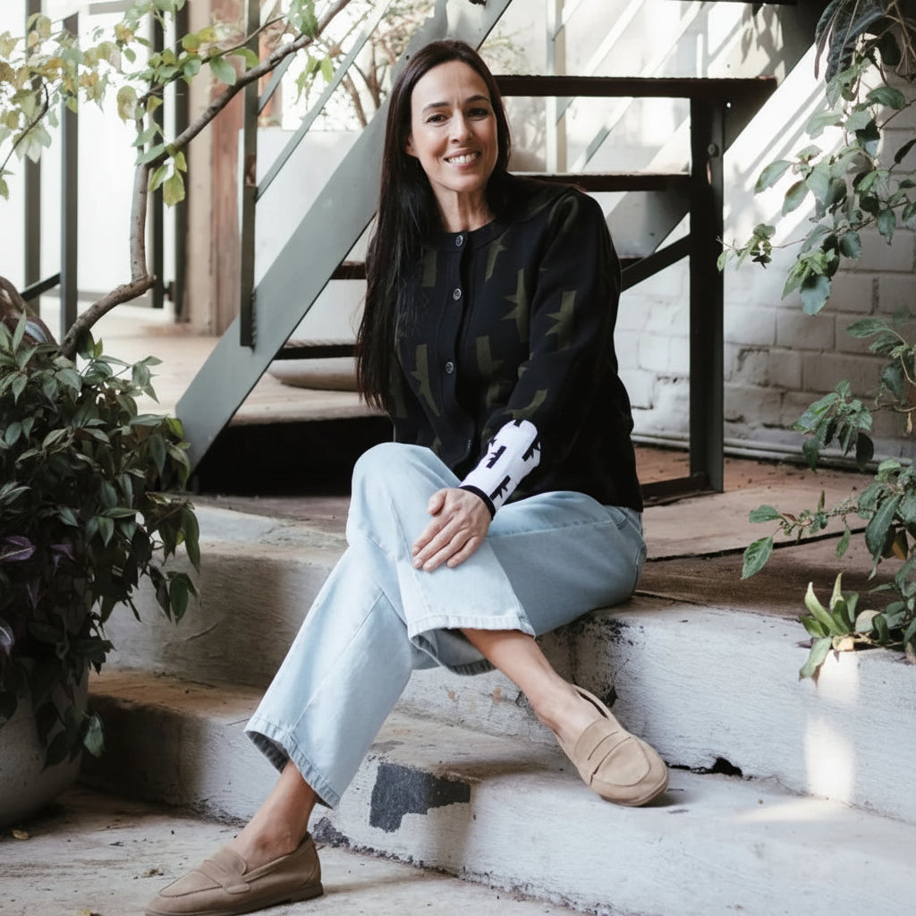 A woman sits on concrete steps in a bright loft-style space with a metal staircase and plants. She is wearing a black long-sleeve top with a green abstract pattern, light-wash wide-leg jeans, and tan slip-on shoes.
