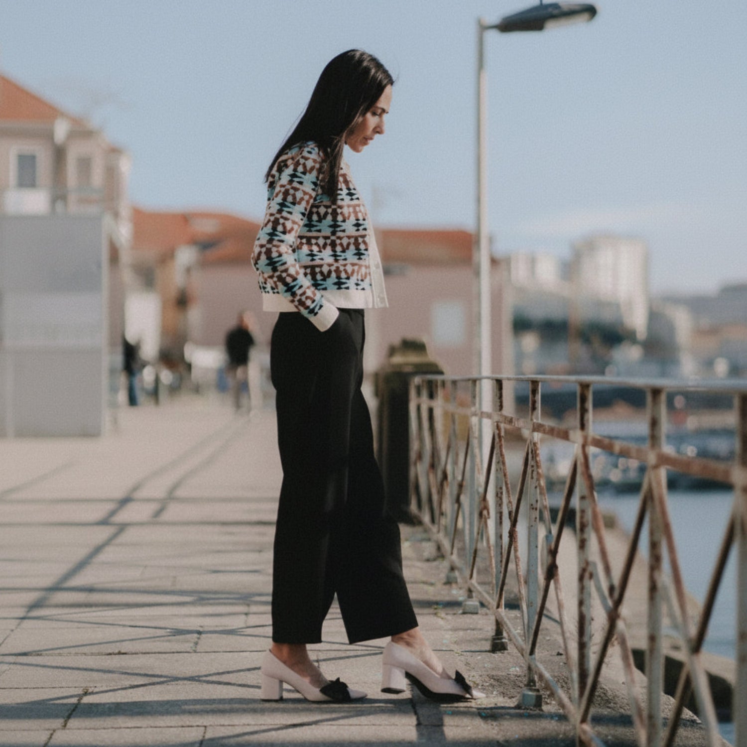 A woman stands on an outdoor walkway wearing a cropped multicolor patterned cardigan, black wide-leg cropped pants, and white block-heel pumps with black bows.