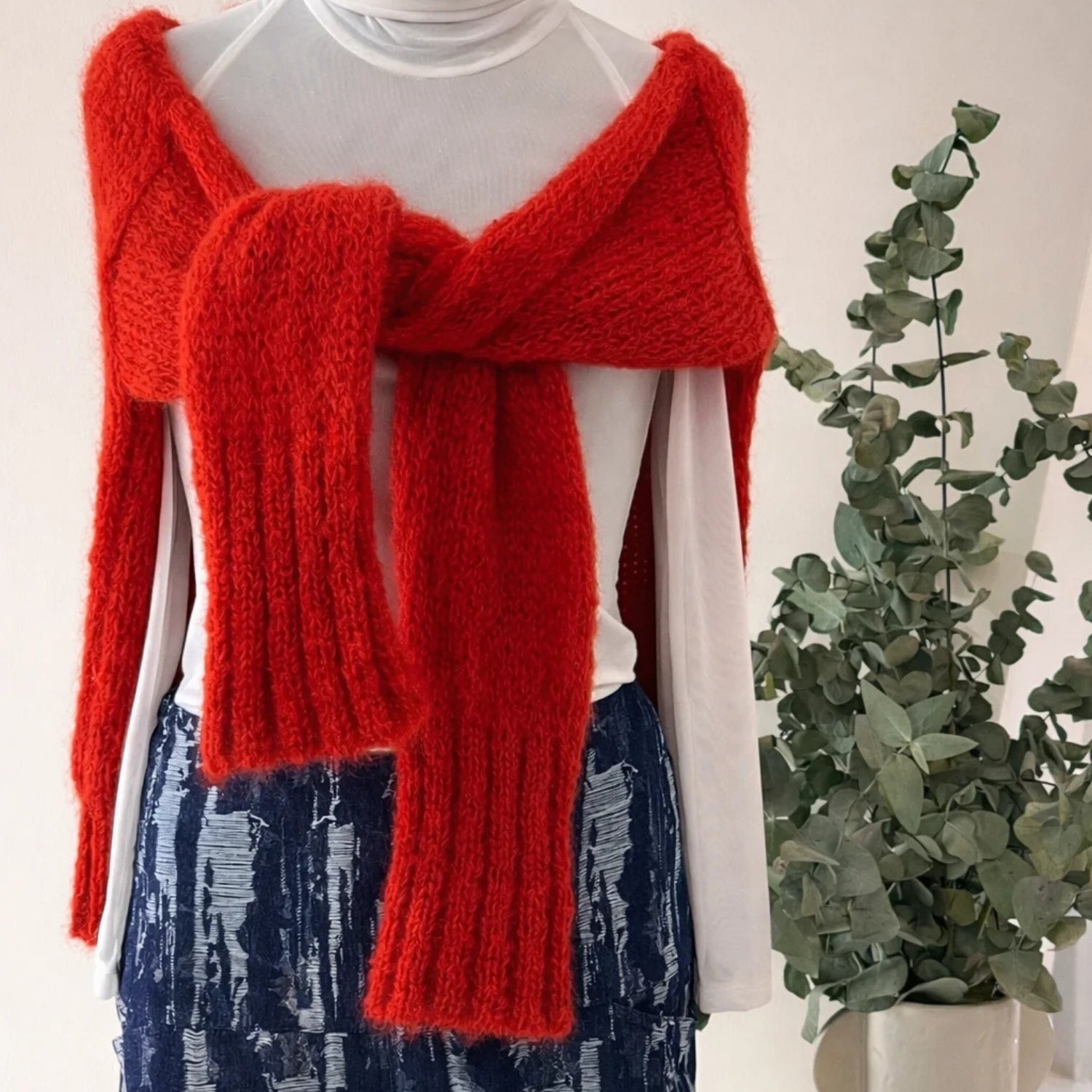 A bright red chunky knit sweater vest styled over a white long-sleeve top and a blue patterned skirt, displayed on a mannequin next to a potted eucalyptus plant.