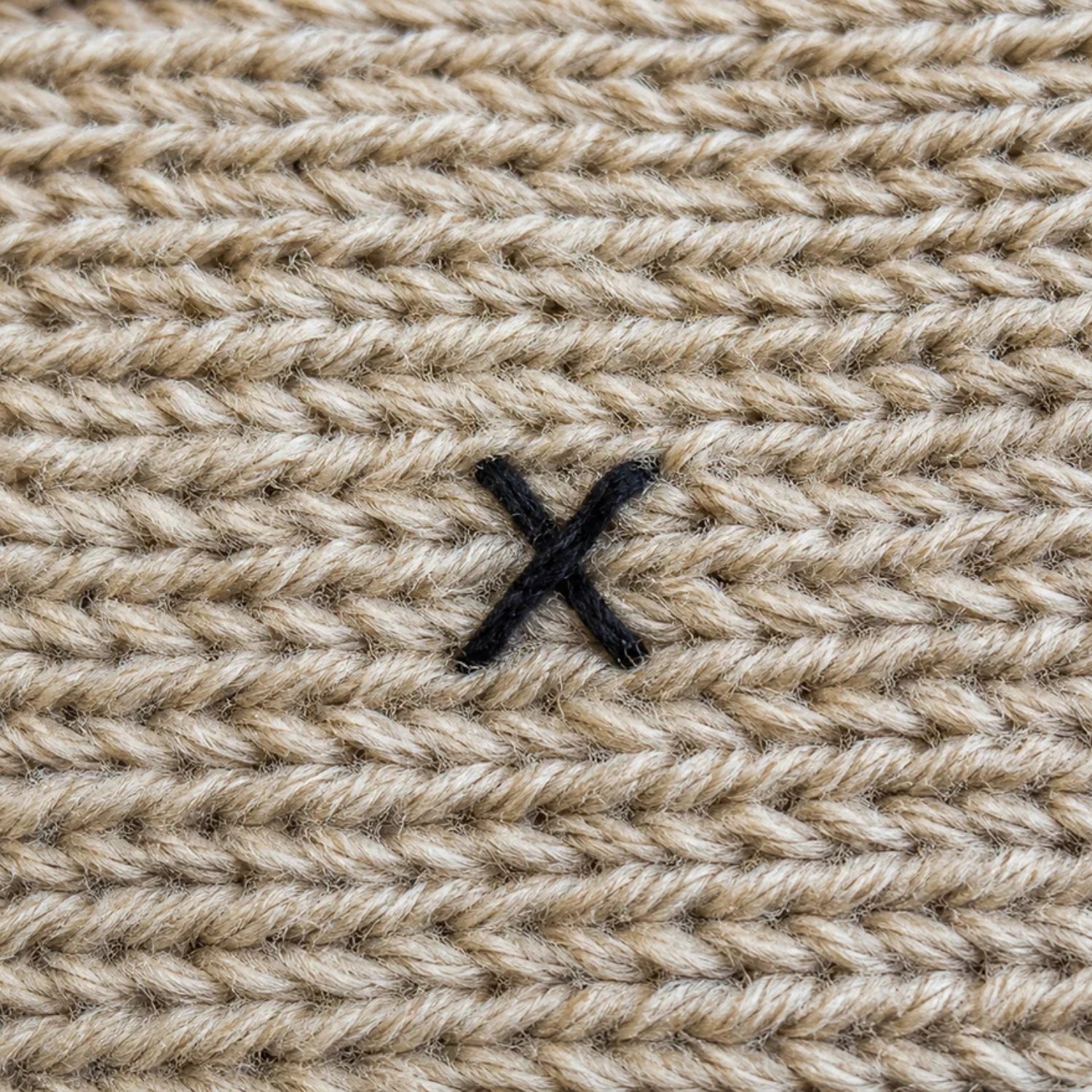 Close-up of a beige textured knit fabric with a small black embroidered X logo in the center.