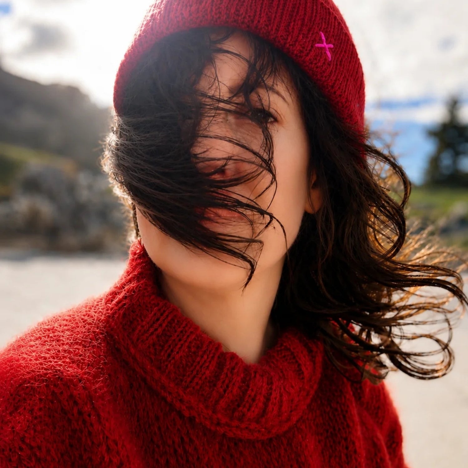 A close-up lifestyle shot of a woman outdoors wearing a vibrant red knit beanie with a small pink cross accent and a matching red knit turtleneck sweater, with her dark hair blowing in the wind.