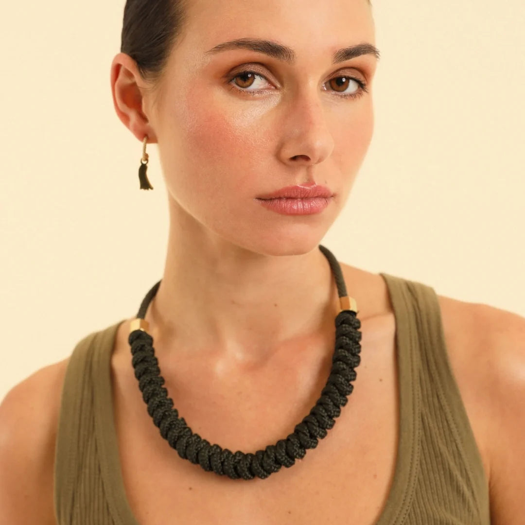 A close-up of a woman wearing a chunky black braided rope necklace with small gold-toned end beads, paired with an olive green ribbed tank top against a warm beige background.