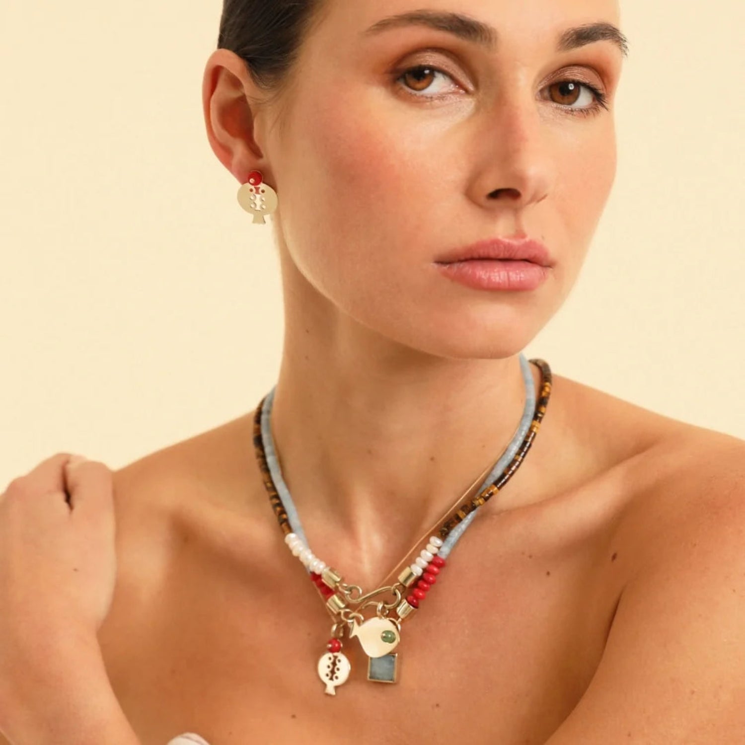 A close-up of a woman wearing a layered beaded necklace with gold charms and a matching gold stud earring, set against a solid light beige background.