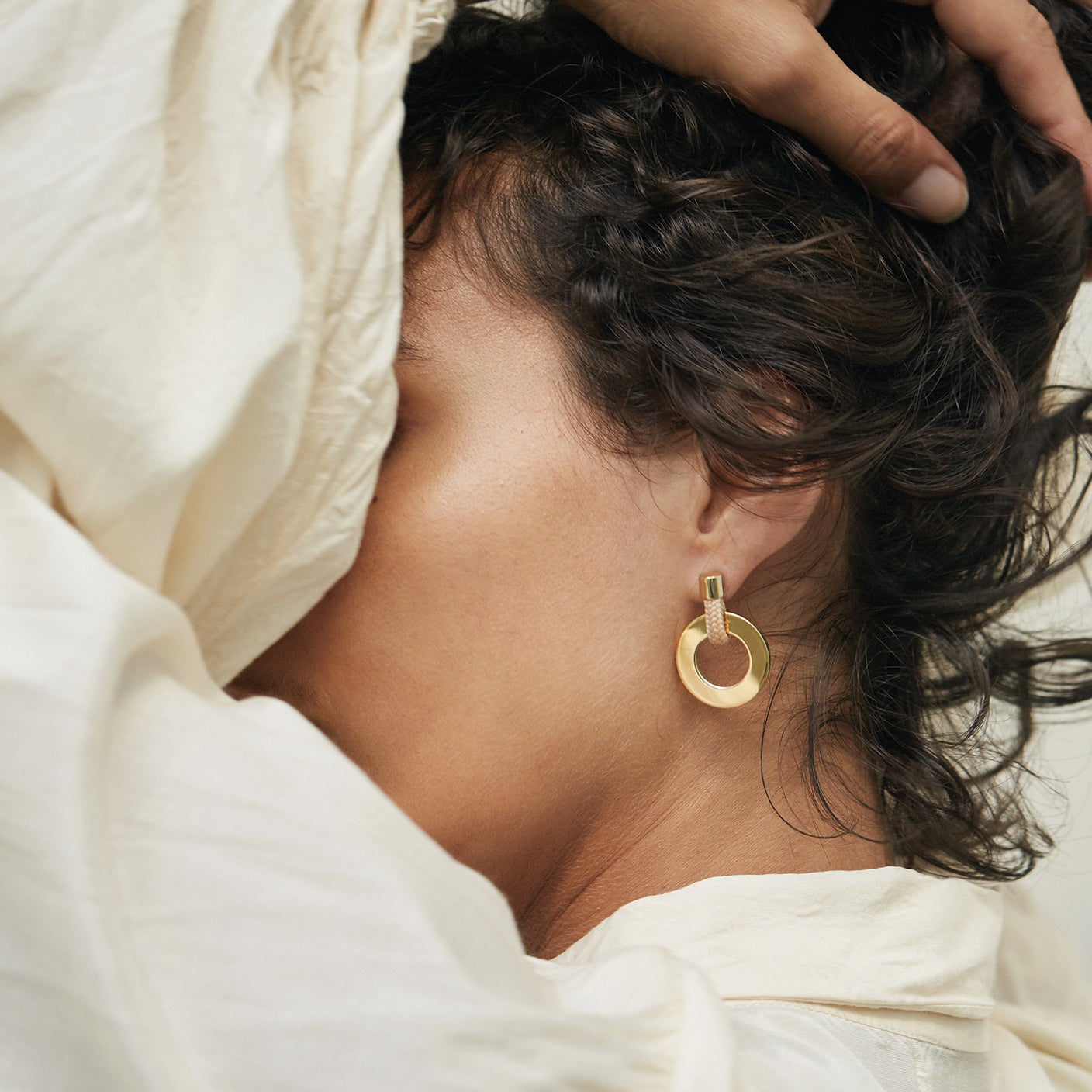 A close-up lifestyle shot of a model wearing a gold circular hoop earring, with her hand running through her dark hair and draped off-white fabric around her head.