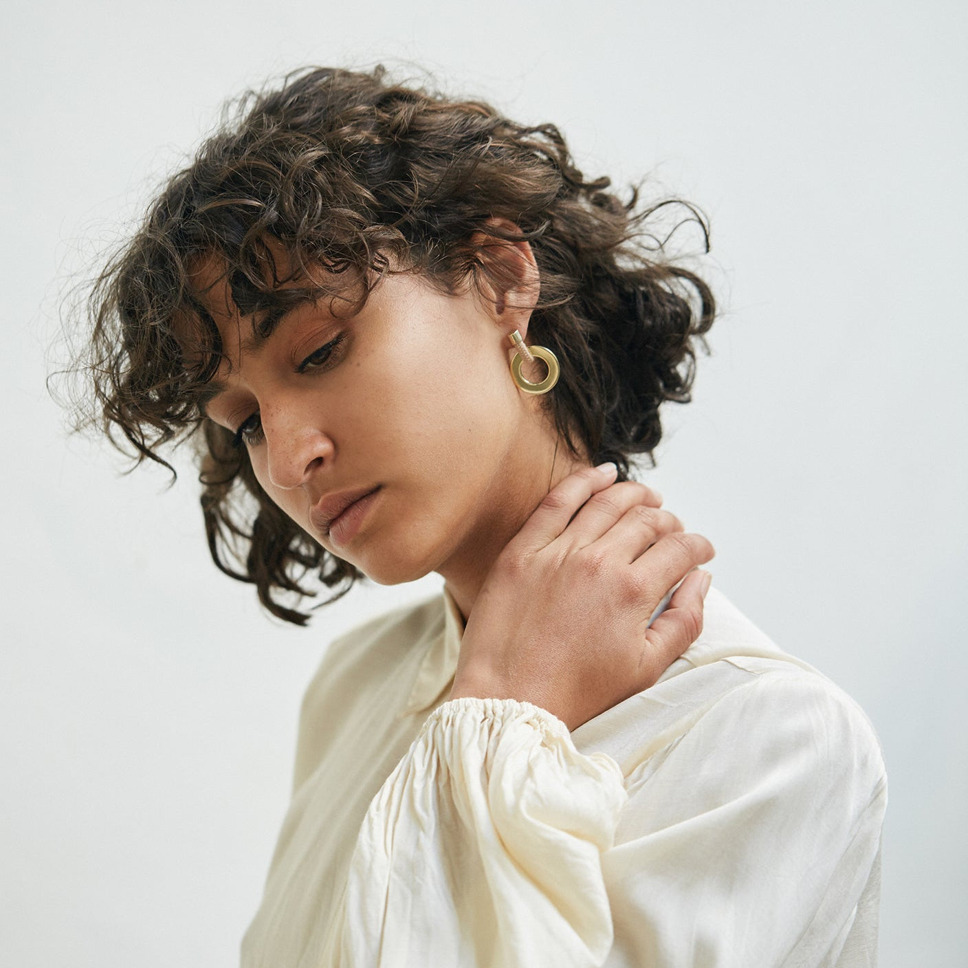 A close-up portrait of a woman with curly dark hair wearing a cream-colored blouse and a gold hoop earring, set against a plain light gray background.