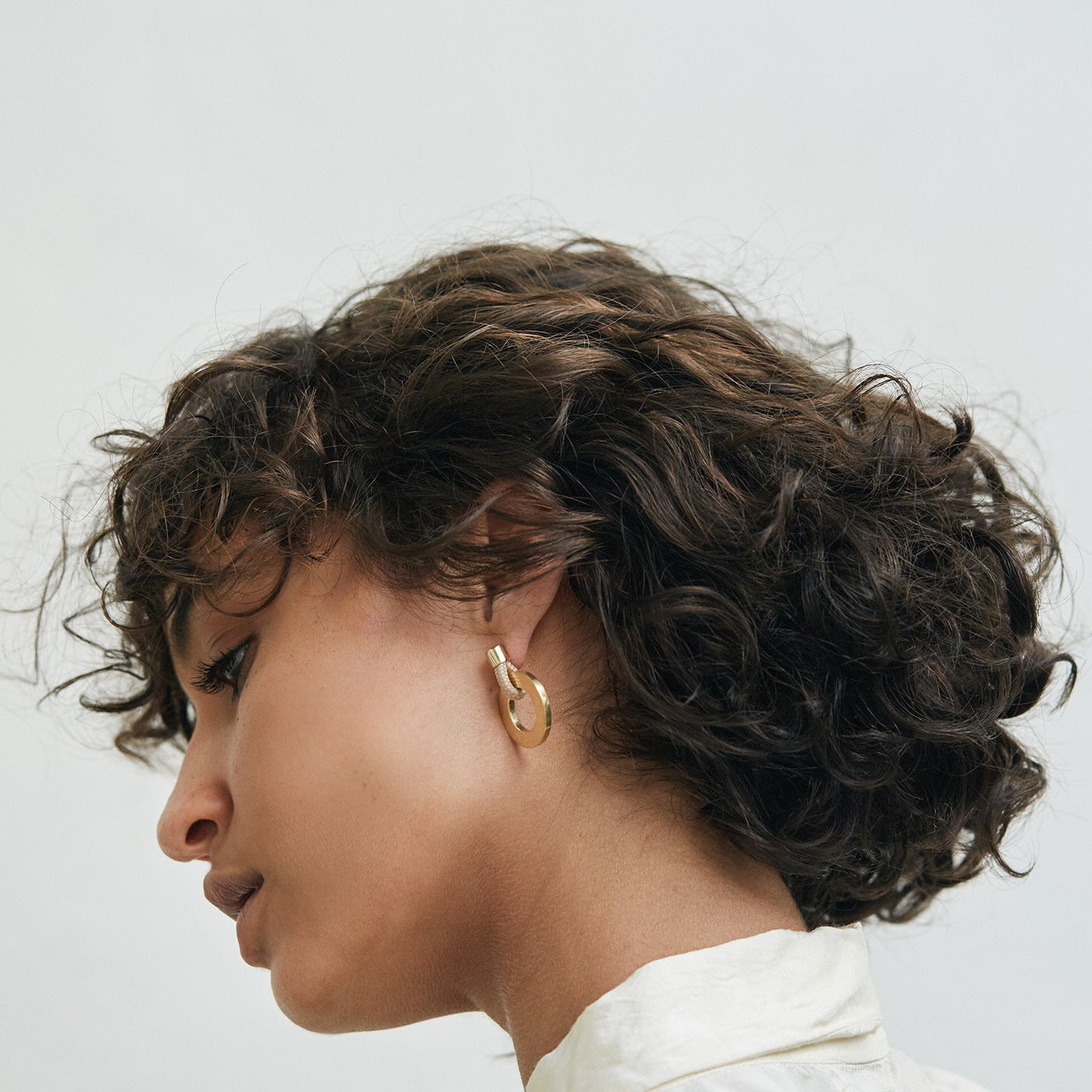 A close-up side profile of a person with short, dark curly hair wearing a gold hoop earring, set against a plain light gray background.